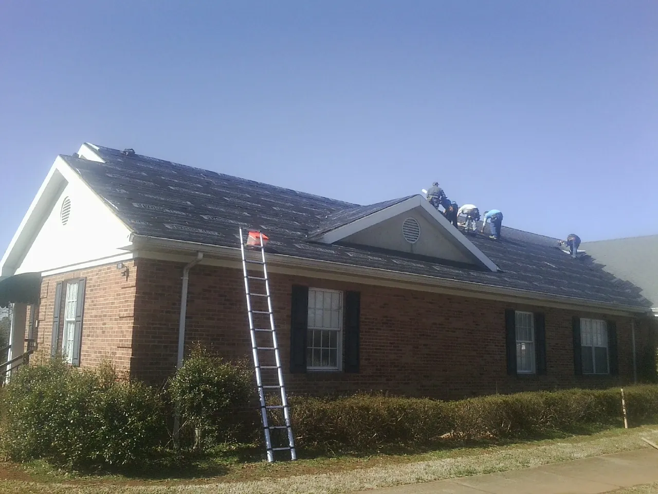 Crew tearing shingles off a brick ranch roof with a ladder set — Emergency Roof Repair work by our crew in Wedgefield