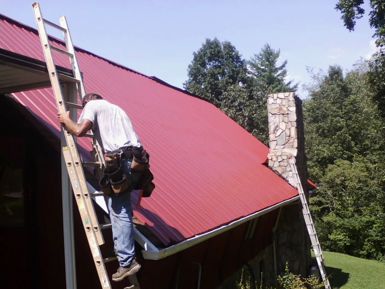 Roofer on a ladder beside a red metal roof and stone chimney — Soffit & Fascia Repair by Roofing Brothers in Wedgefield